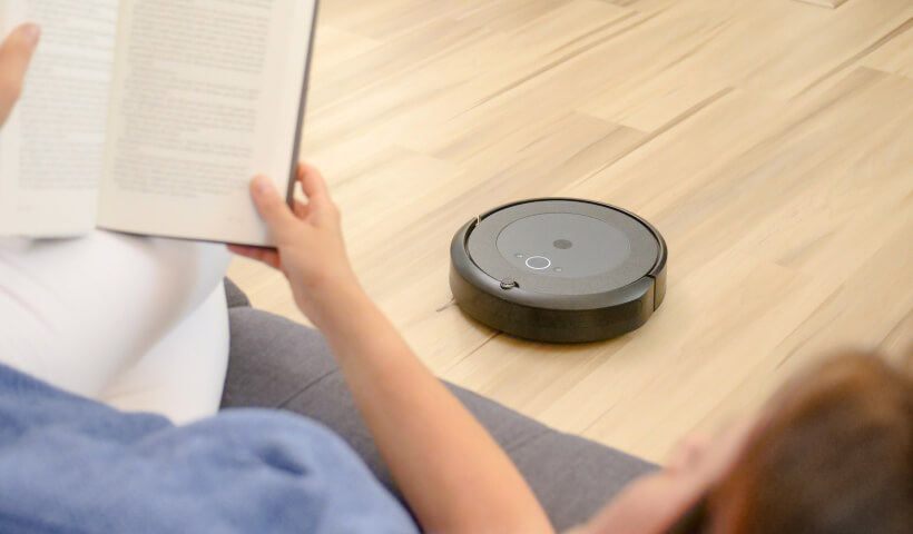 Photo d'une femme qui lit un livre dans son canapé pendant qu'un aspirateur robot passe l'aspirateur dans son salon.