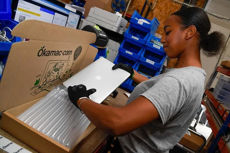 Opératrice en train d'emballer soigneusement un MacBook reconditionné dans le packaging Okamac